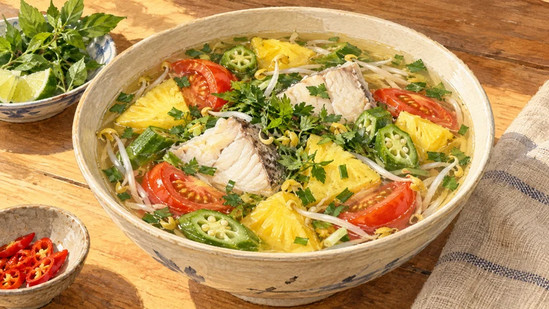 Illustration of white fish, pineapple, tomato, okra, bean sprouts and herbs in a bowl of clear golden broth.