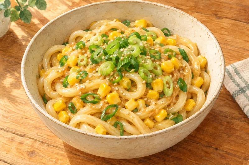Watercolor illustration of thick udon noodles coated in miso butter with yellow corn, green scallions and sesame in a cream bowl.