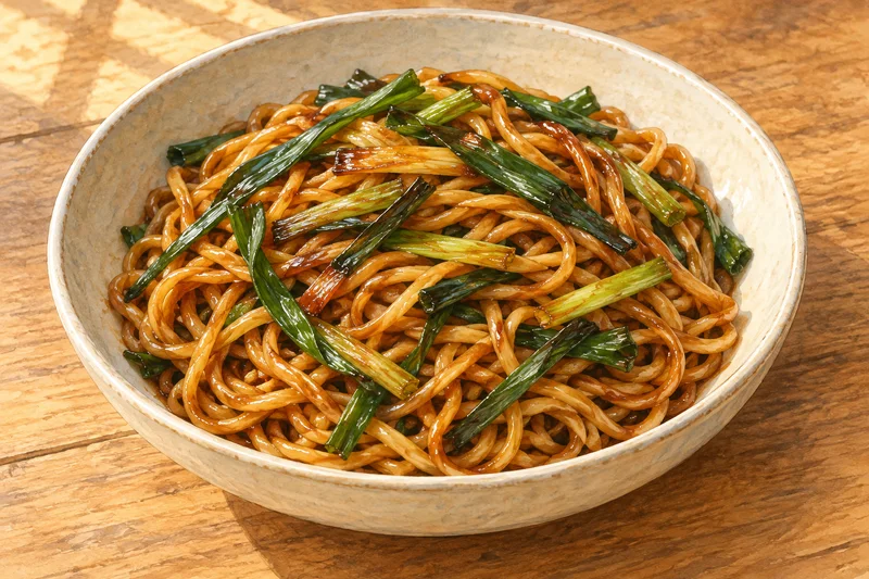 Watercolor illustration of glossy amber wheat noodles with long browned scallion pieces in a cream ceramic bowl.