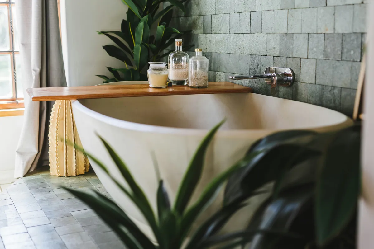 Lush green houseplants beside a freestanding bathtub in a modern bathroom with green tile walls and a wooden bath tray