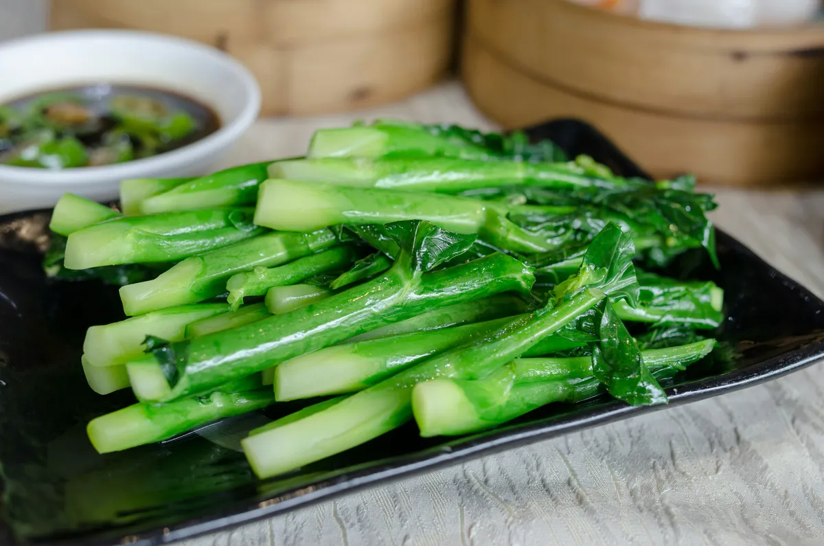 Blanched choy sum with bright green stems and leaves arranged on a black plate with a small bowl of soy dipping sauce and bamboo steamers in the background