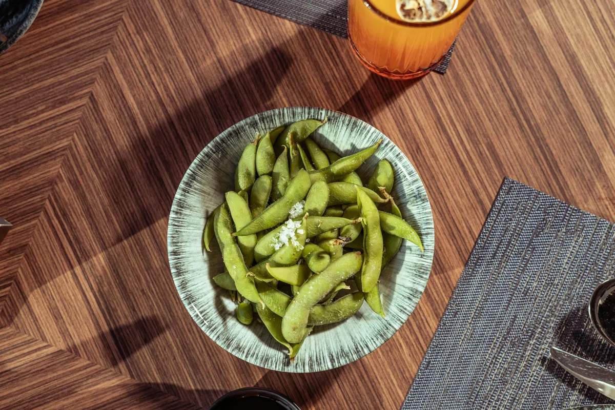 Aerial view of fresh edamame pods sprinkled with coarse salt in a ceramic bowl on a wooden table